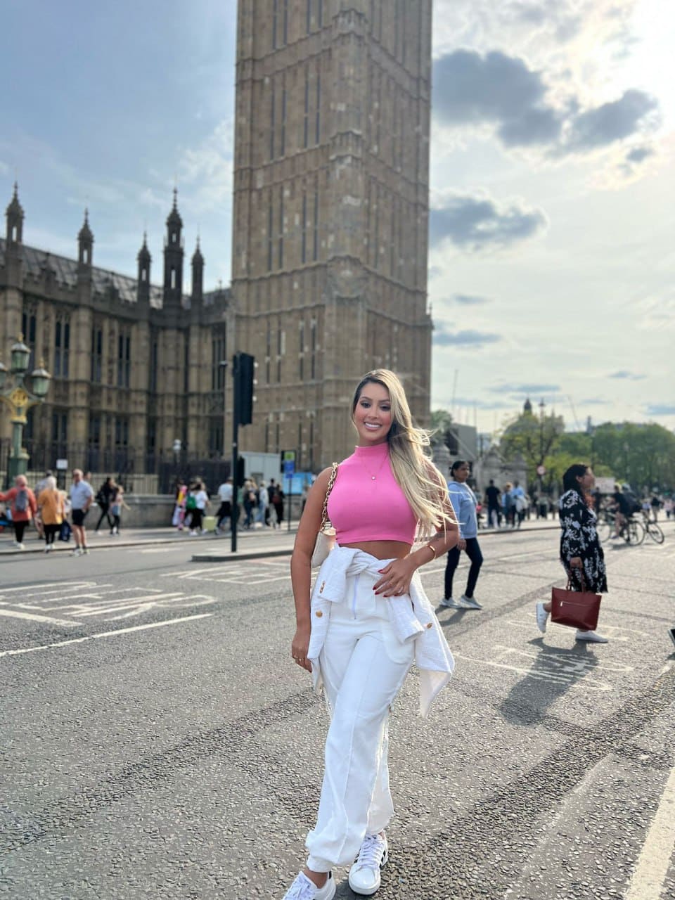 Dani stands on a city street in a pink cropped turtleneck, white trousers, and sneakers, with a stone tower behind her.