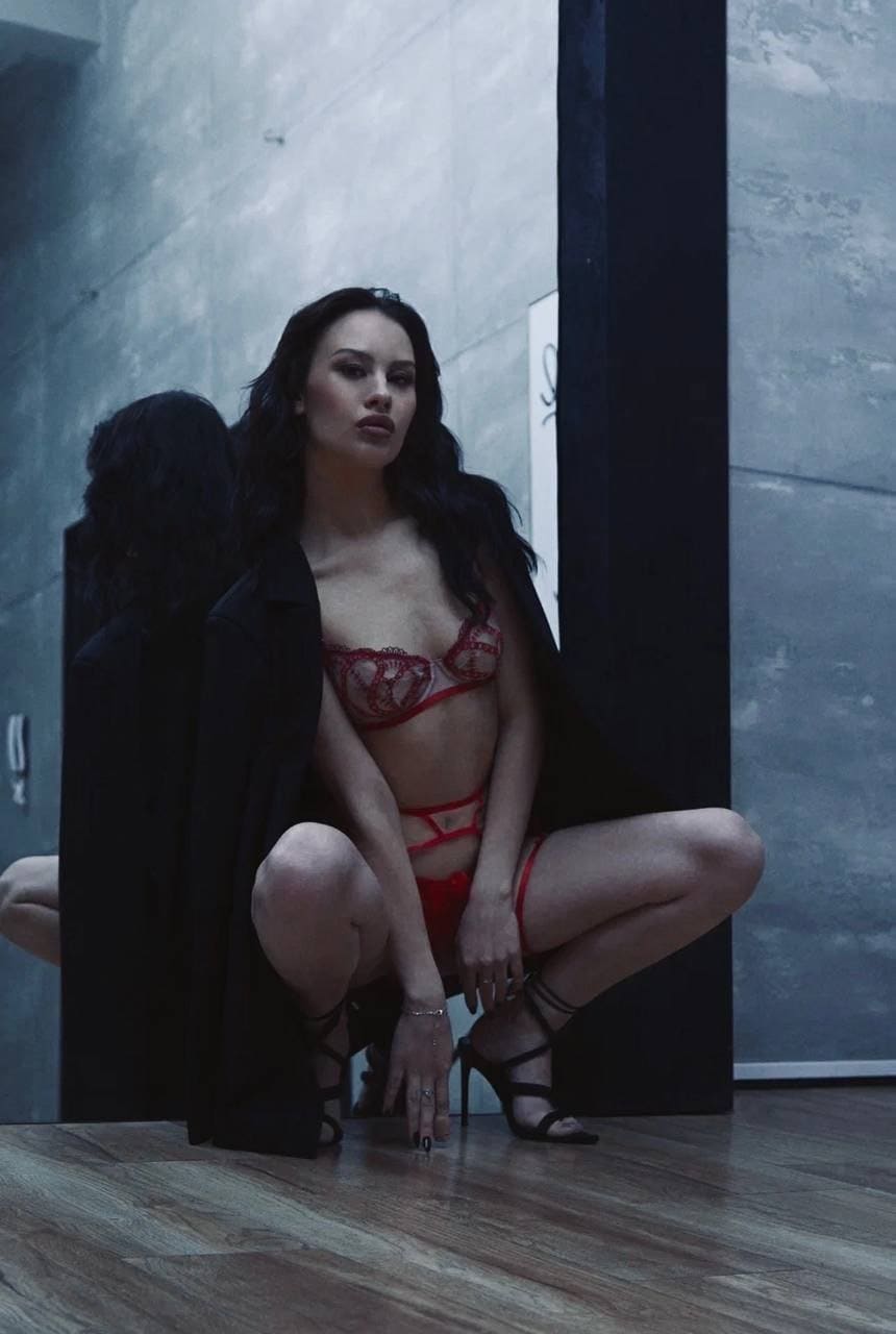 A woman crouches on a wooden floor in red lace and heels, wearing an open black jacket against a gray wall.