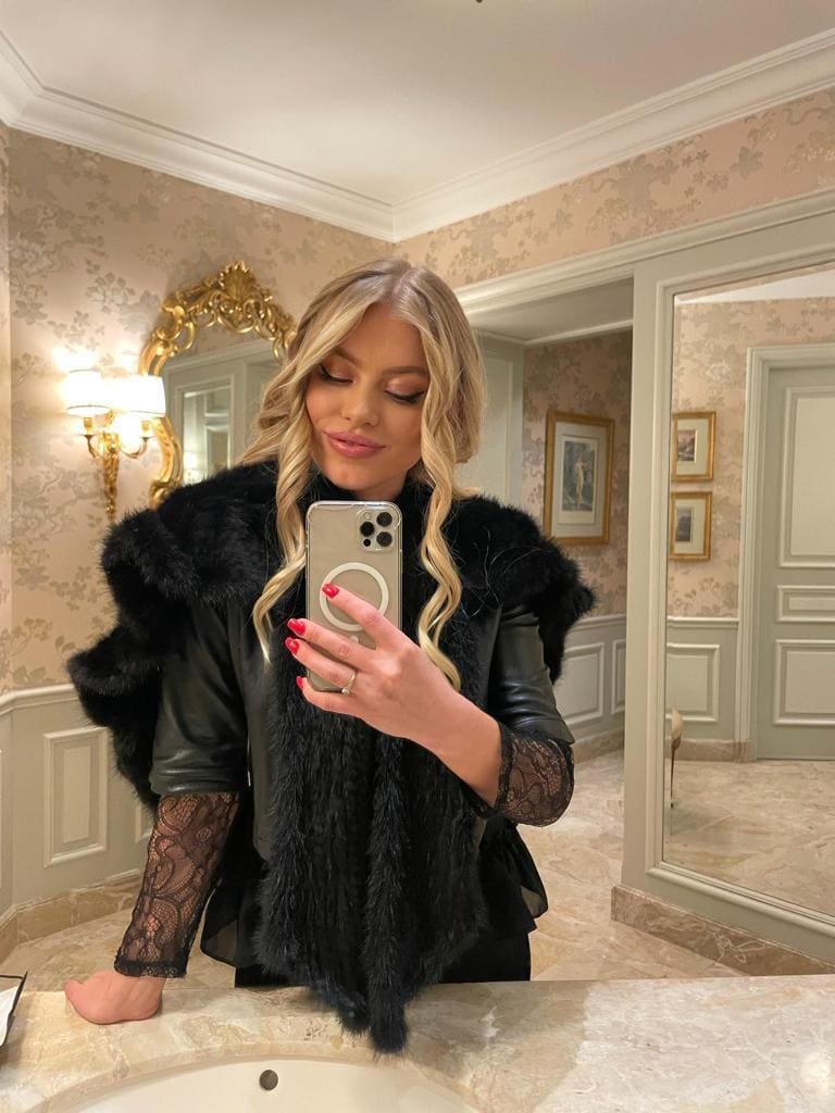 A mirror selfie shows a woman in a black leather jacket with fur collar and lace top, holding a phone in a bathroom.