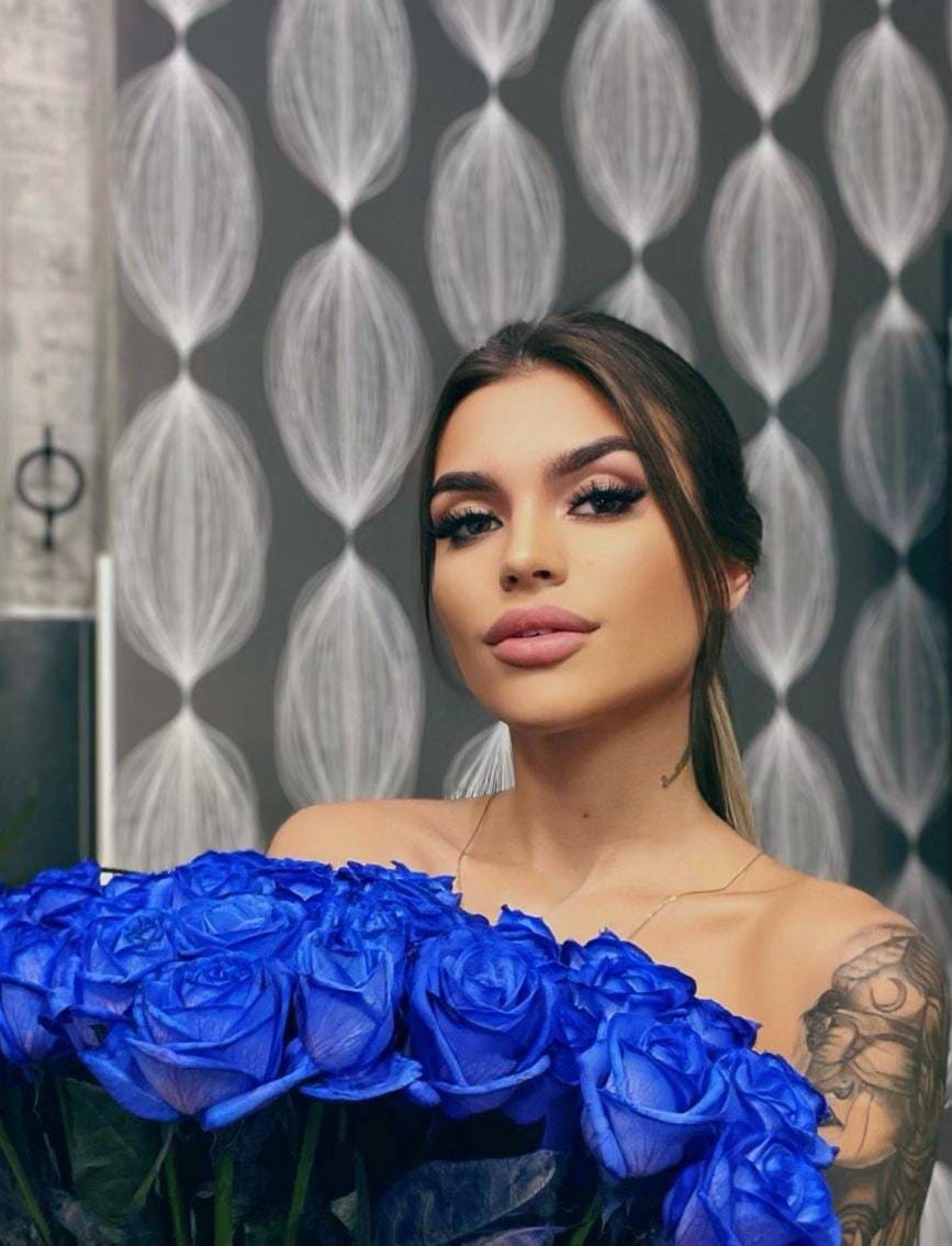 A woman with a shoulder tattoo holds blue roses against a patterned wall, wearing an off-shoulder top and thin necklace.