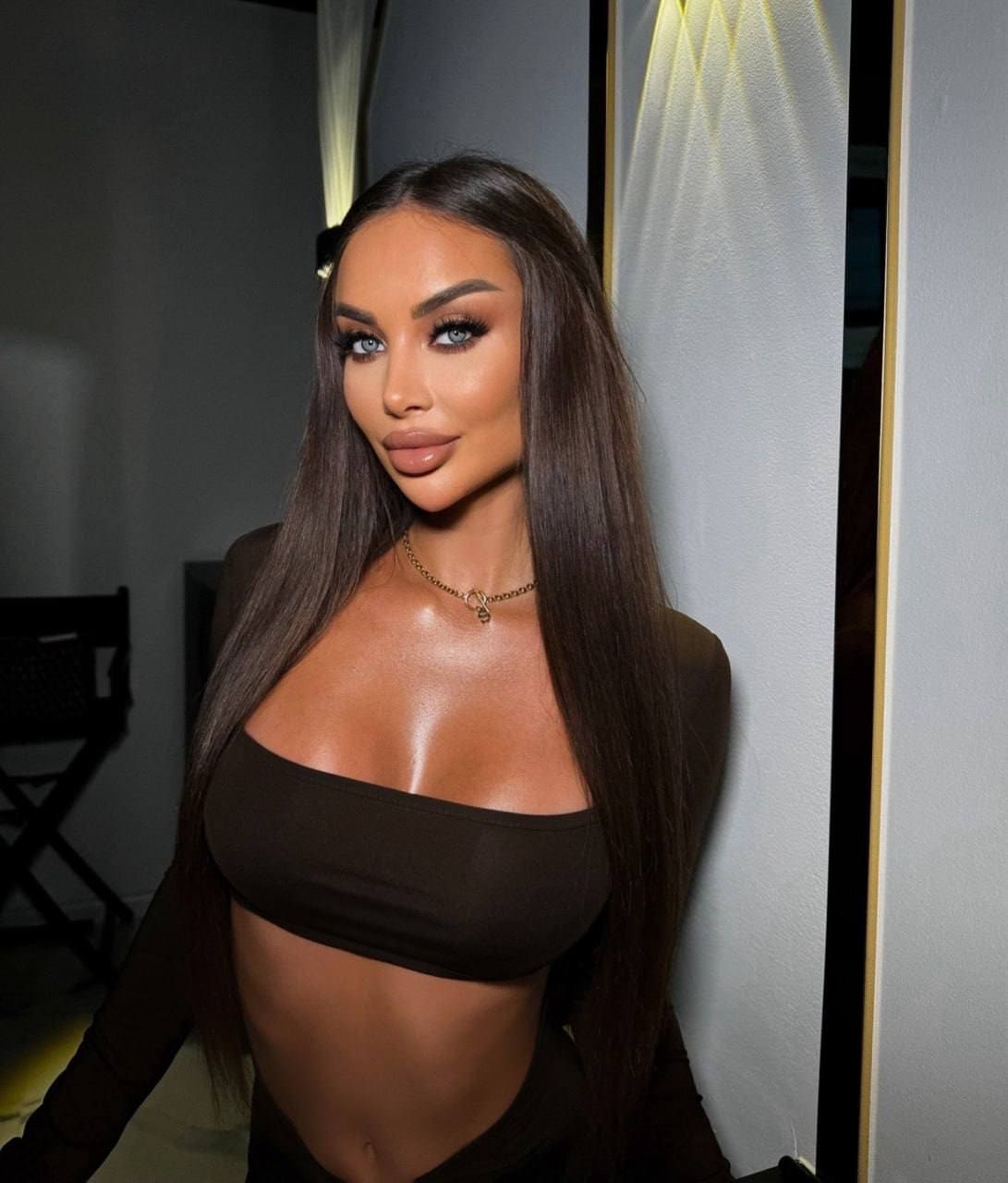 Standing facing the camera in a dark strapless crop top and matching bottoms, with a thin chain necklace, against a light wall and dark chair.