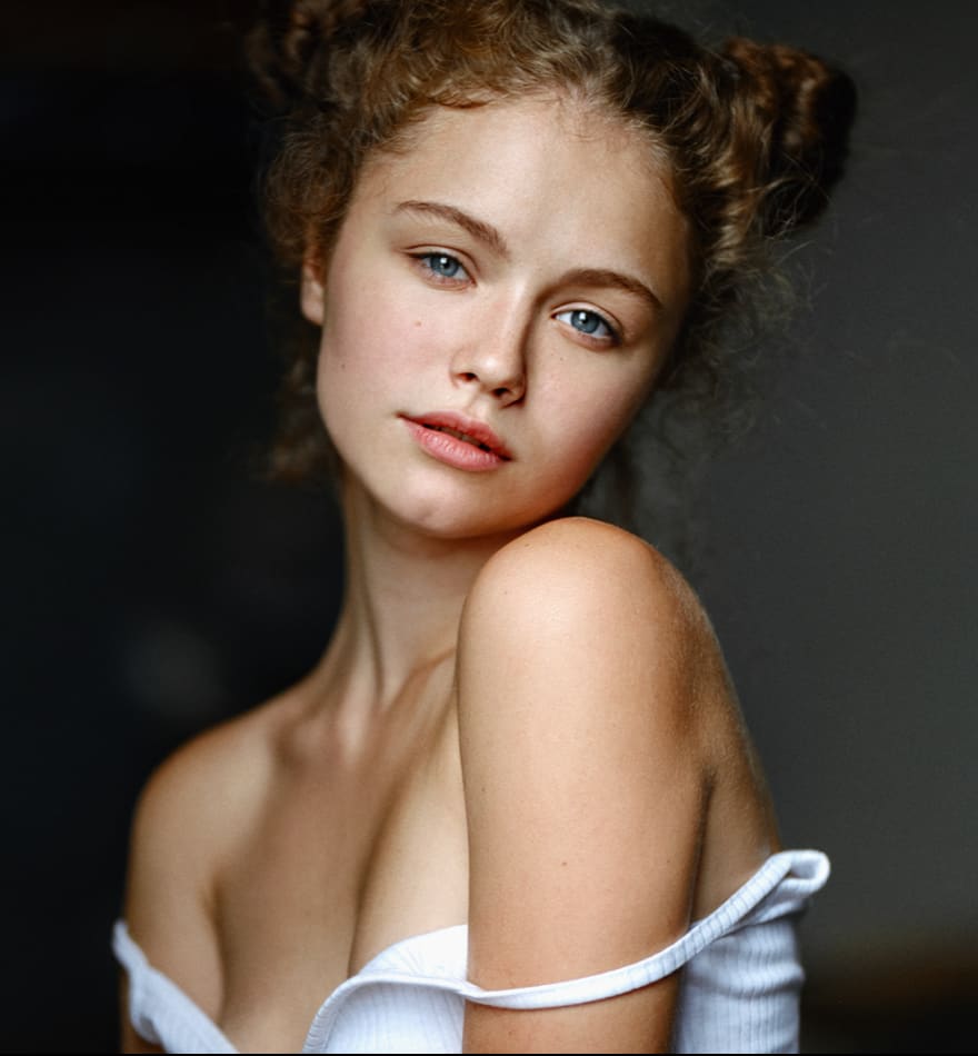 Ophelia with curly brown hair in two buns tilts her head, wearing a white off-the-shoulder top against a dark background.