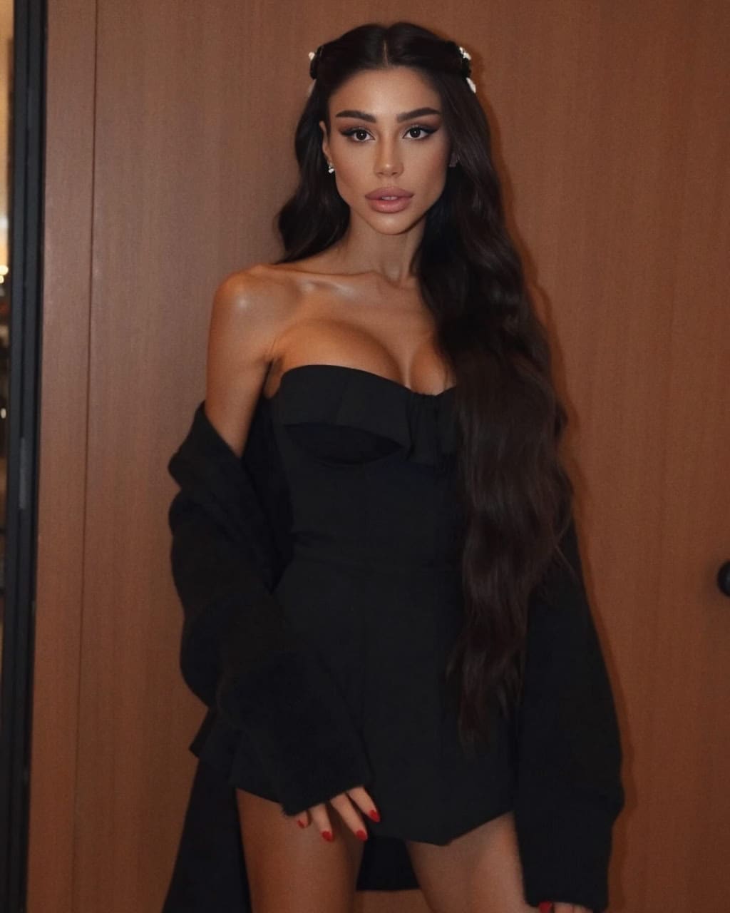 Olivia stands by a wooden wall in a black strapless mini dress and jacket draped off her shoulders, hand near her thigh.