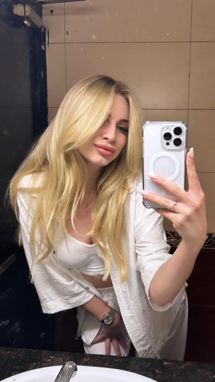 A blonde woman takes a mirror selfie in a bathroom wearing a white bralette and open cardigan, holding a silver smartphone.