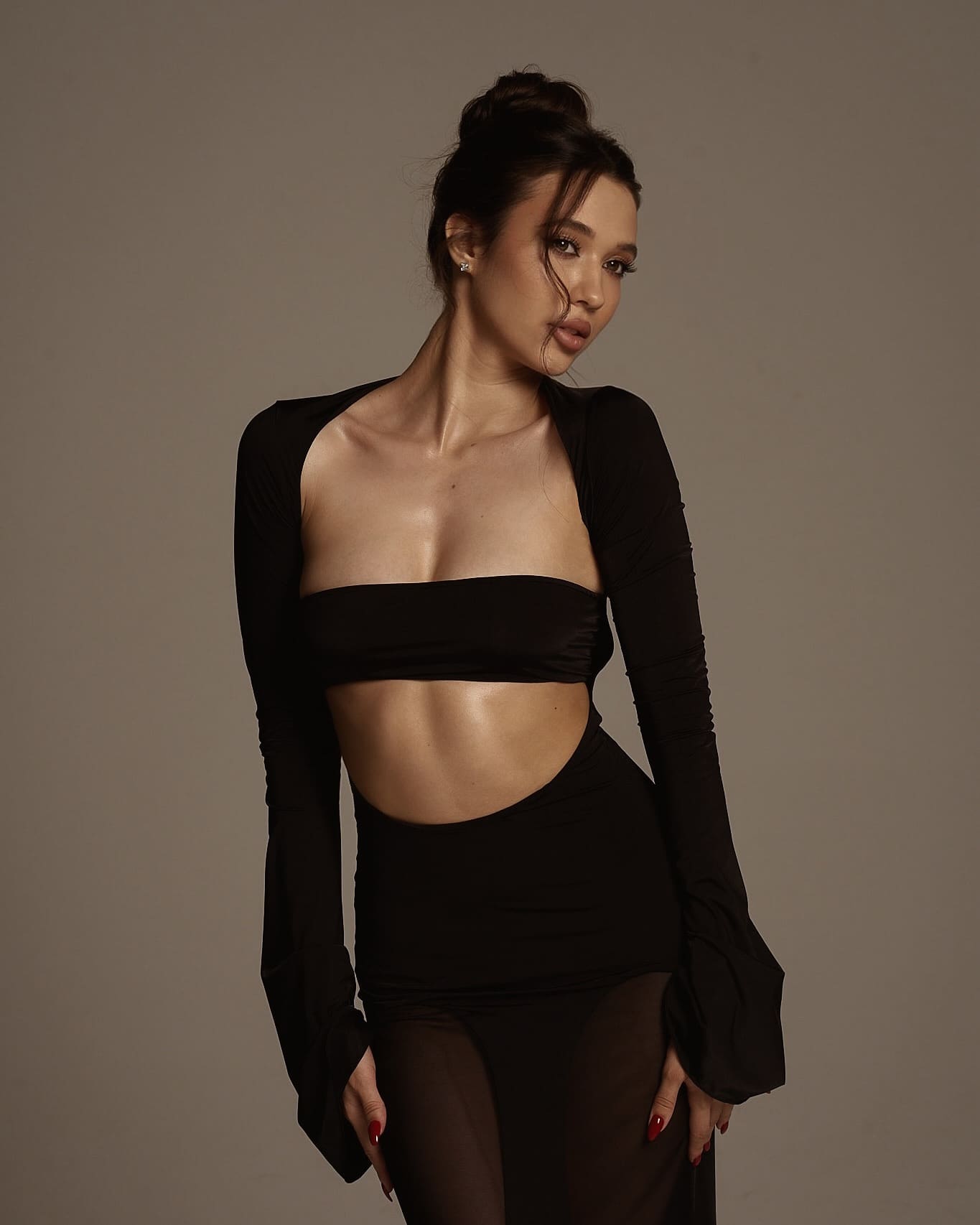 Mirana stands against a gray backdrop in a black cutout top and sheer tights, hands at her sides with red nails.