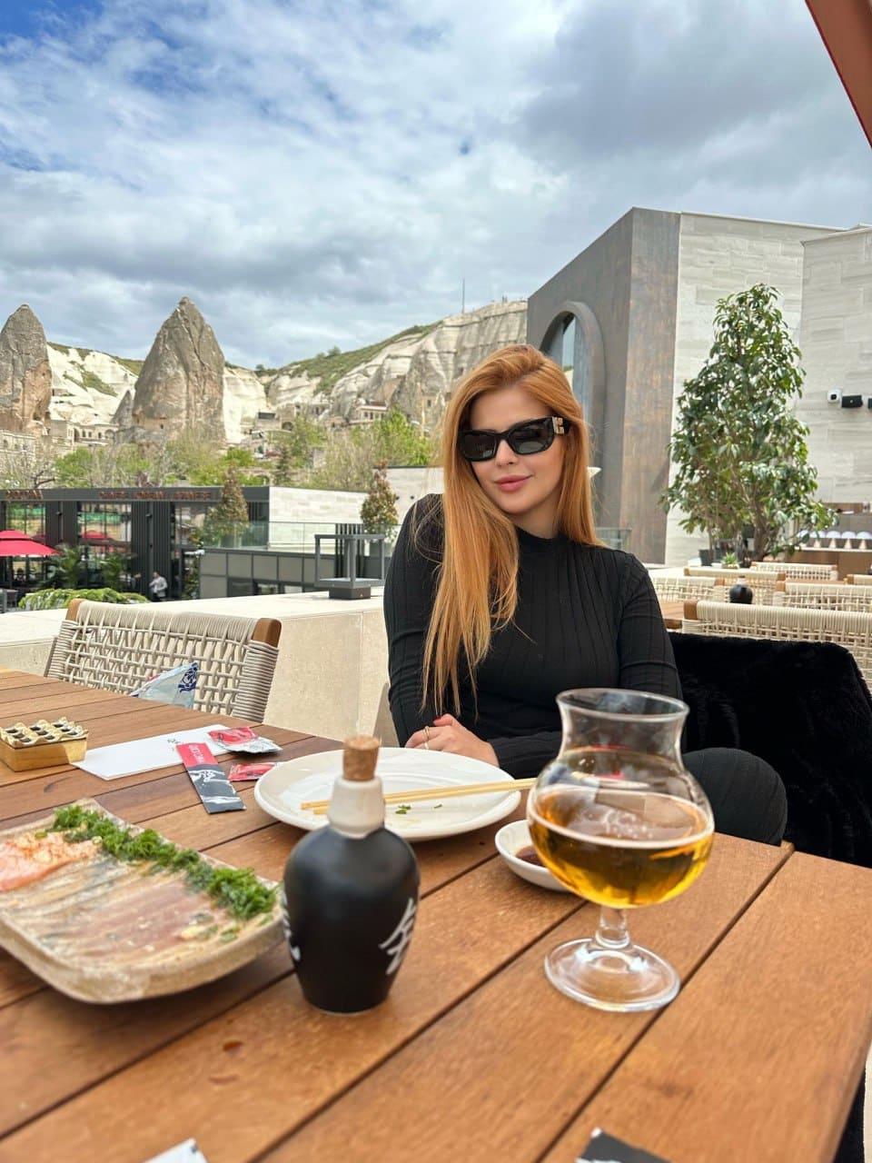 Woman in sunglasses and a black top sits at an outdoor table with food and drinks, rocky hills and a building in the background.