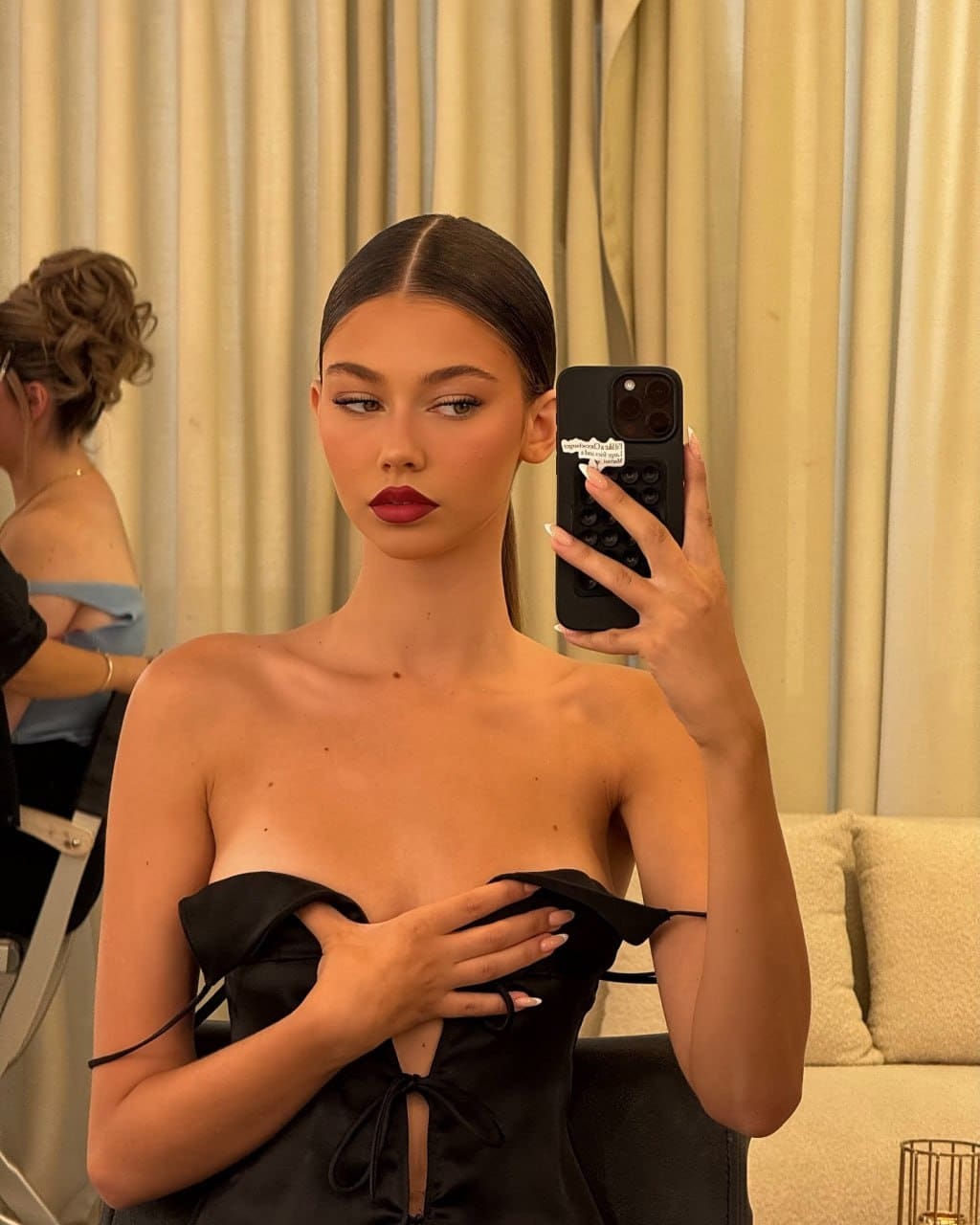 A woman with a sleek ponytail takes a mirror selfie in a black strapless top with side ties, standing near beige curtains and a sofa.