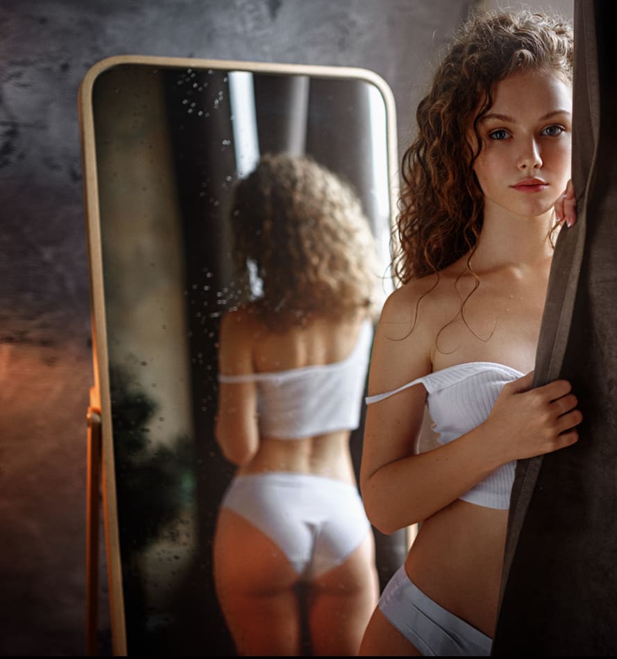 Woman in white strapless top and underwear stands by a mirror, holding a dark curtain, with her back visible in the reflection.