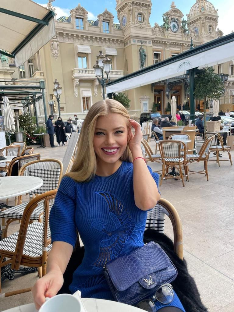 Anna sits at an outdoor cafe table in a blue ribbed sweater with a bird design, smiling with a hand in her hair.