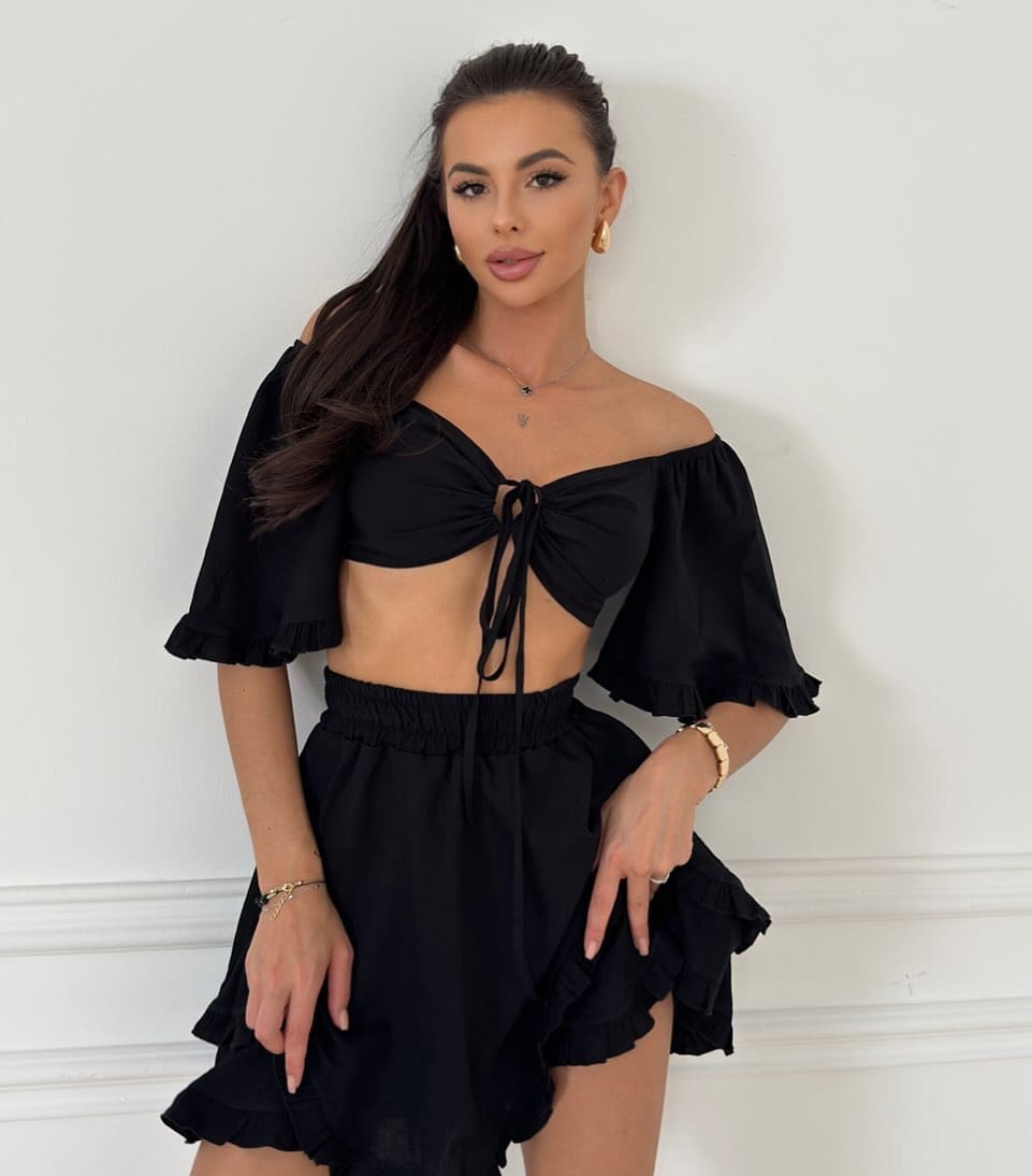 Rinna stands against a white paneled wall in a black off-the-shoulder crop top and ruffled mini skirt, hands near her hips.