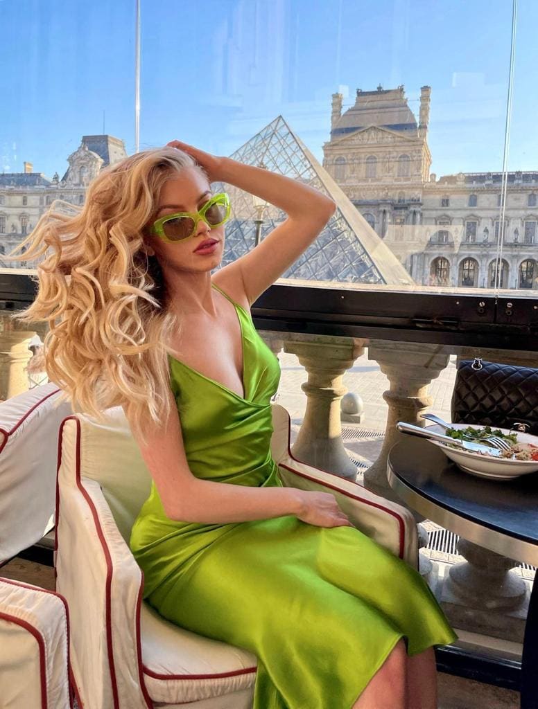 A woman in a green satin dress and sunglasses sits in a white chair before a window overlooking a glass pyramid.