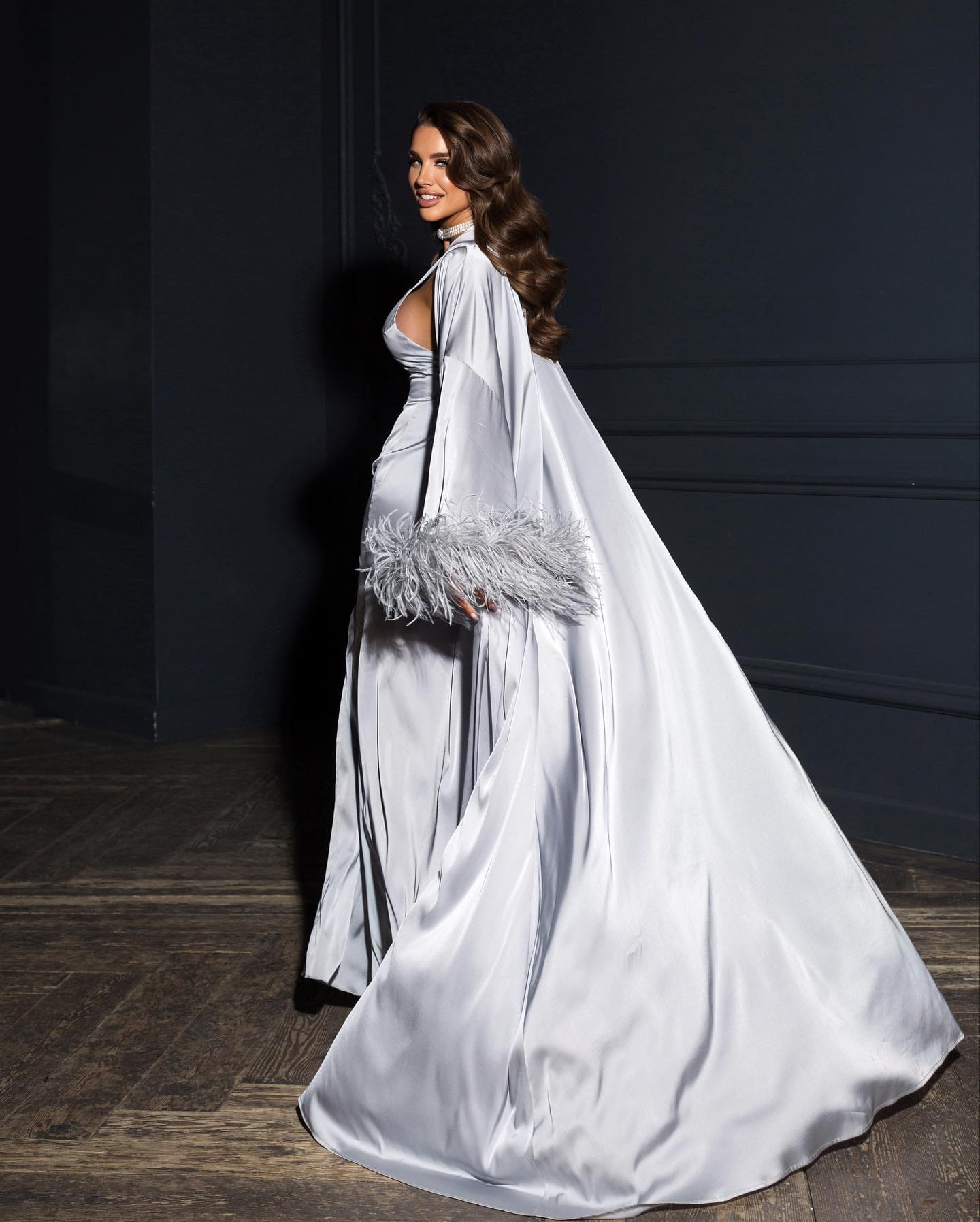 A woman in profile wears a floor-length silver satin gown with an open back and cape, holding a feathered clutch.
