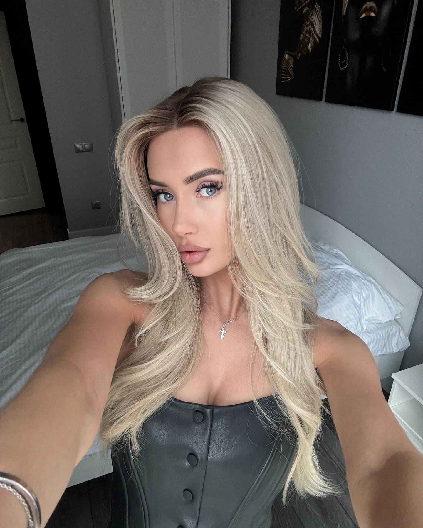 Rita takes a bedroom selfie wearing a black buttoned leather top and cross necklace, with long wavy platinum hair and bed art behind.