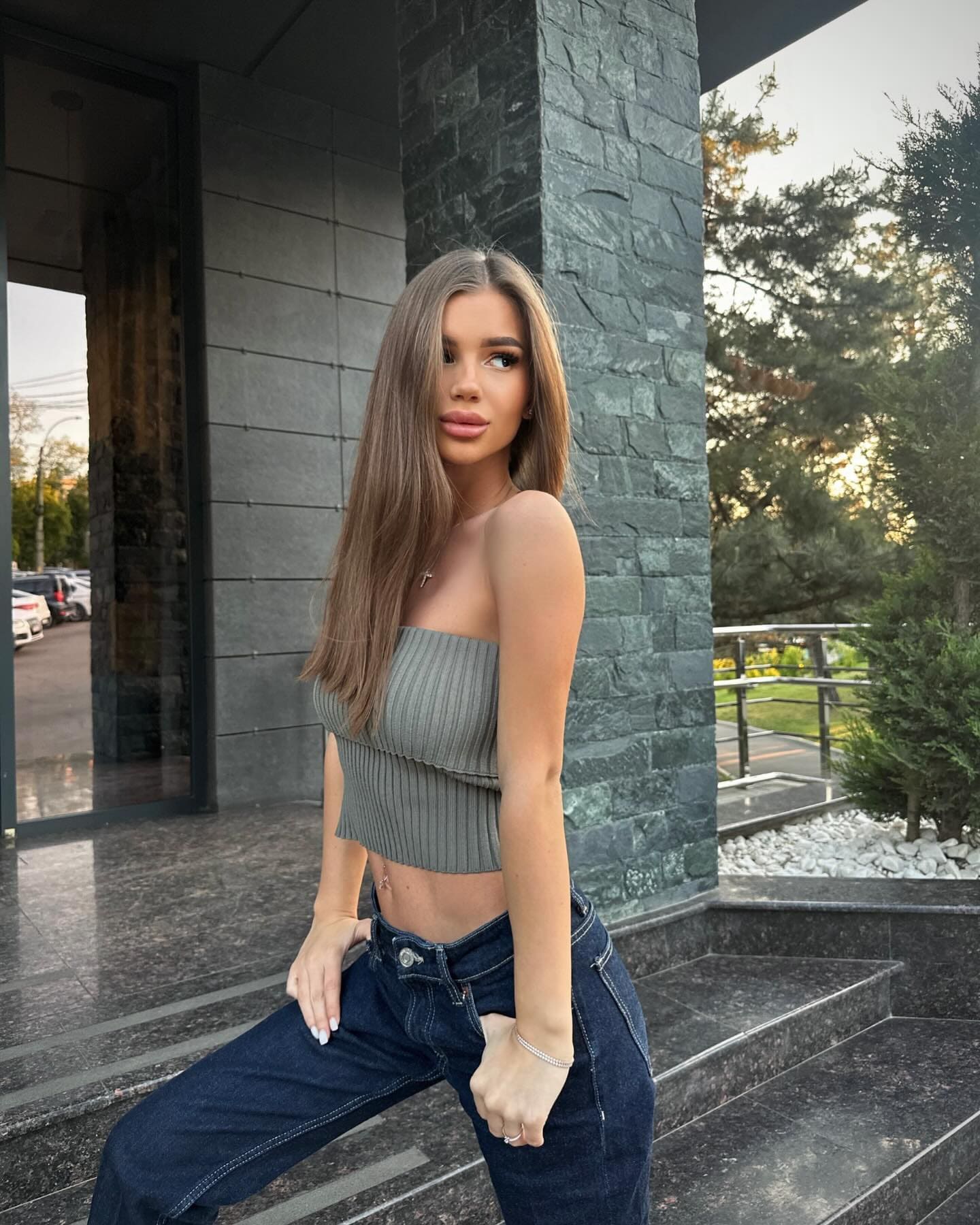 Angel stands on stone steps in a grey strapless crop top and dark blue jeans, one hand on her thigh.