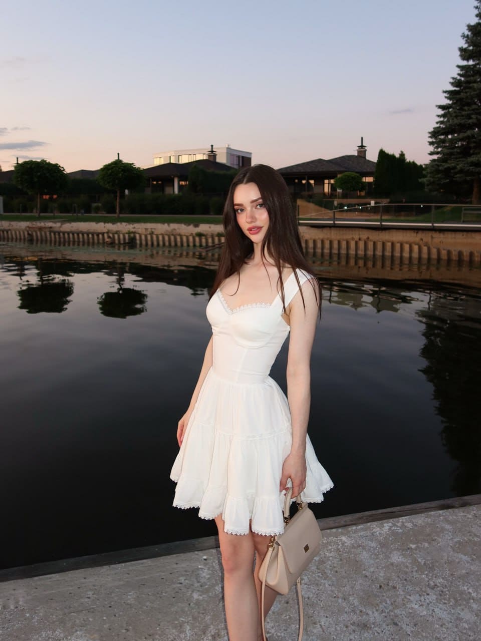 At dusk, a woman in a white lace-trimmed dress stands on a concrete ledge by water, holding a beige handbag and facing the camera.