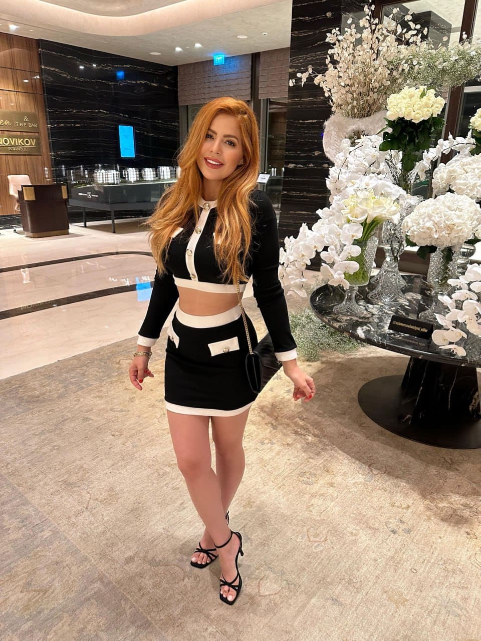 Woman stands in a hotel lobby in a black and white two-piece outfit and black heels, with white flowers on a table to her right.