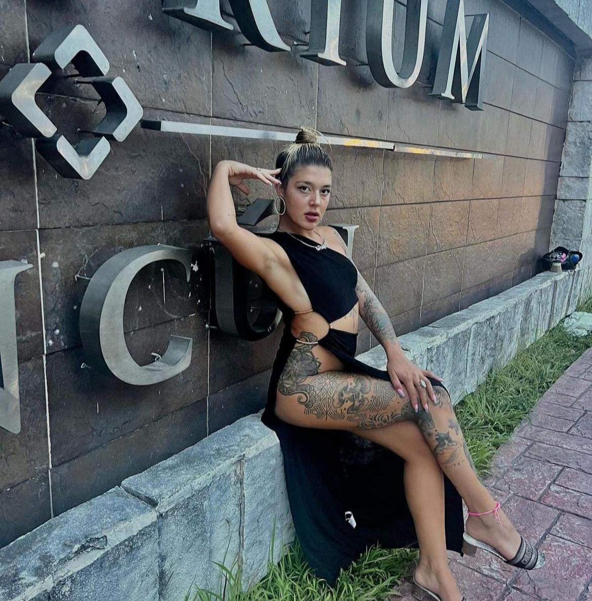 Delfina sits on a stone ledge in a black cut-out dress and strappy sandals, posing with one hand near her head against a dark tiled wall.