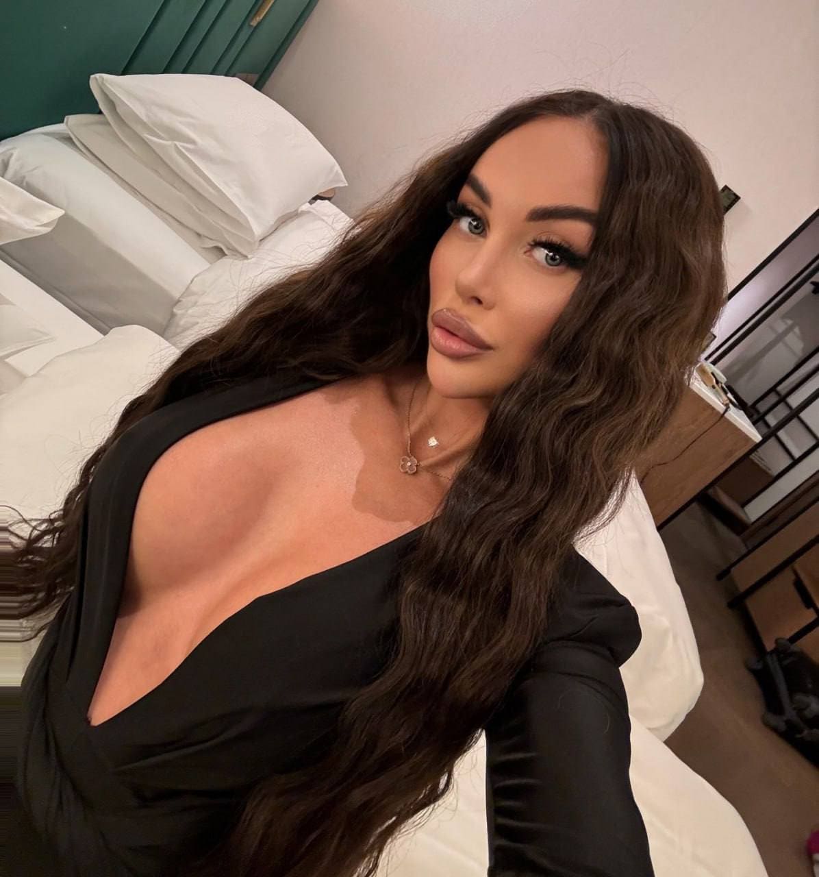 Seated on a white bed wearing a black long-sleeved top with a deep plunging neckline and a thin pendant necklace.