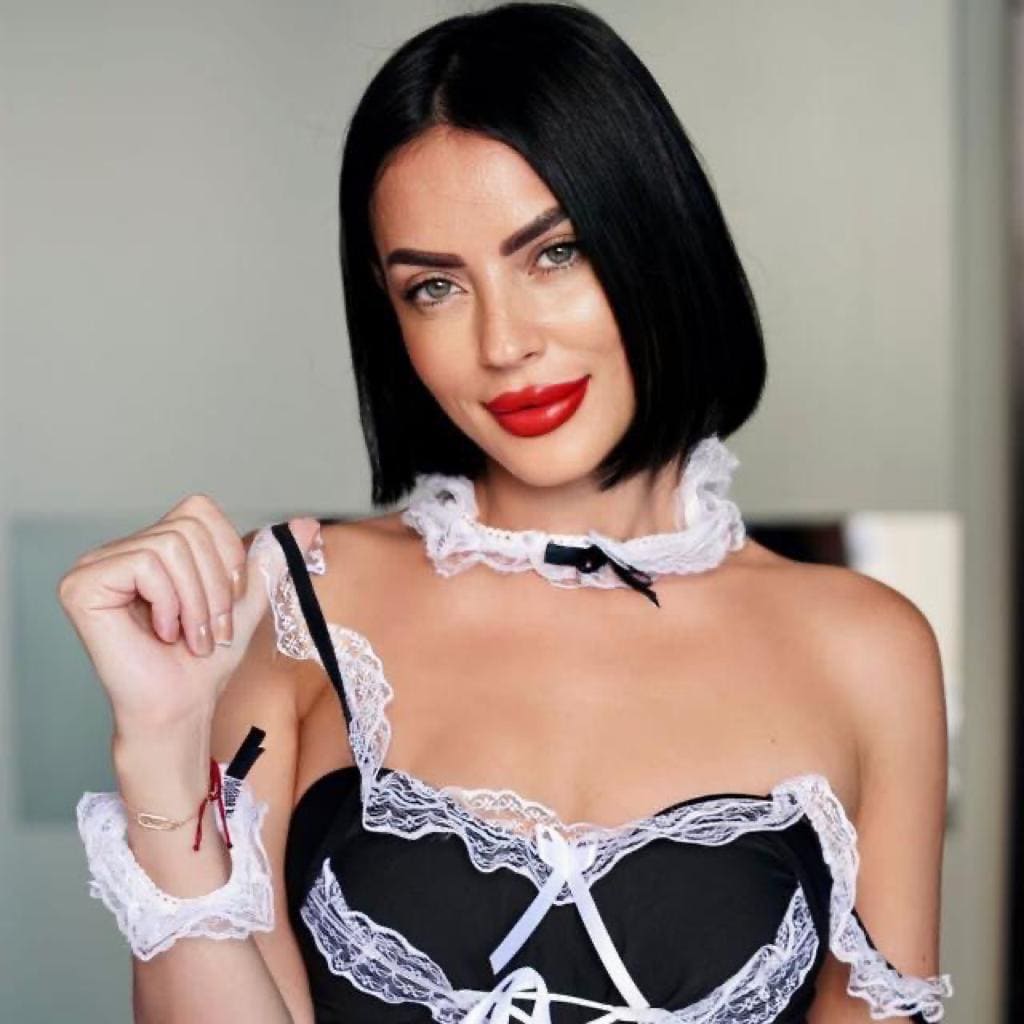 Alice wears a black lace bodice with white ribbon lacing and choker, holding a strap against a plain light wall.