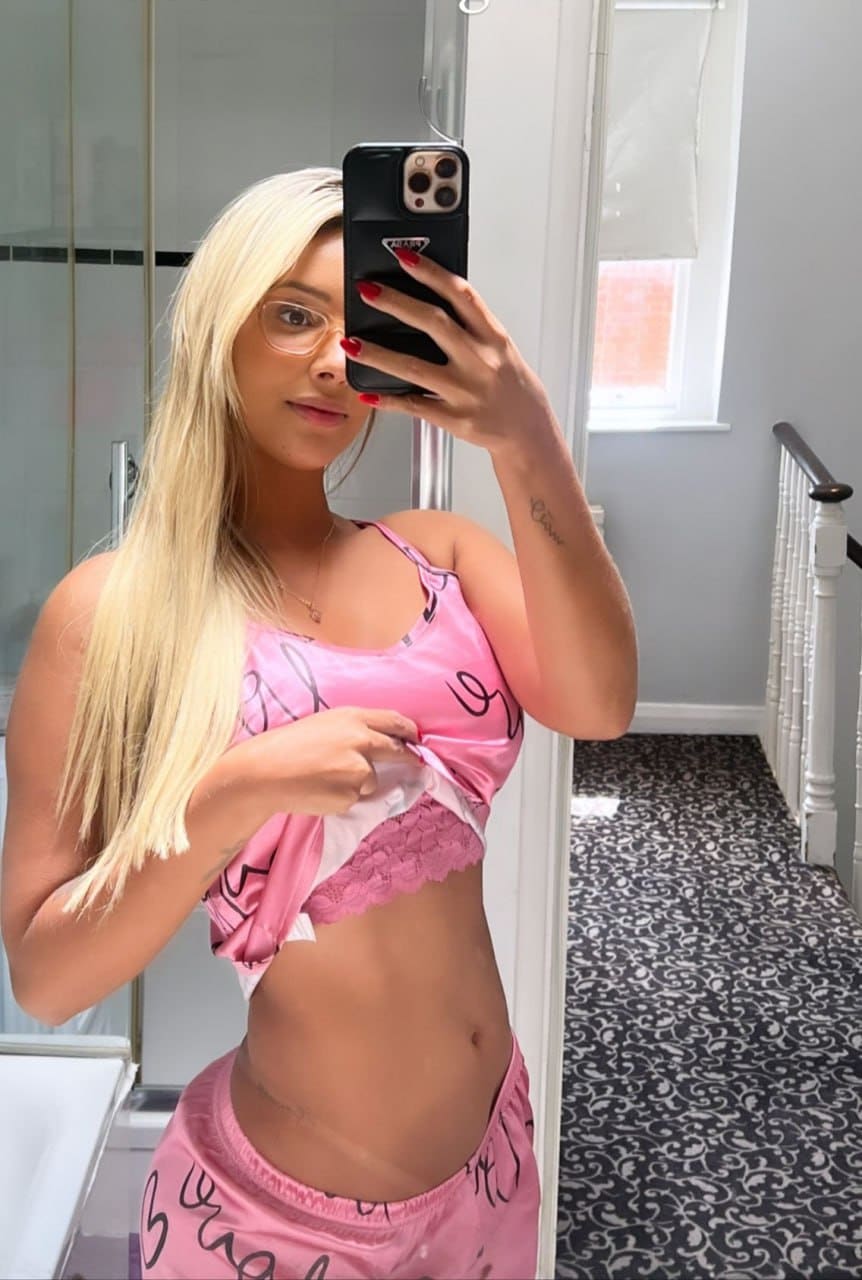 A woman in glasses takes a mirror selfie in a pink satin outfit, lifting her top to show lace underwear in a hallway.