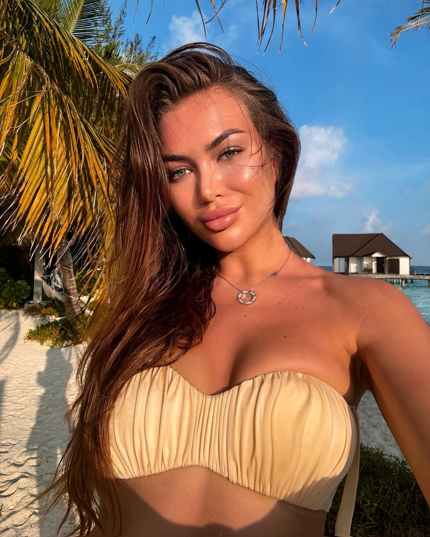 A woman in a strapless cream top stands on a sandy beach, with palm fronds and overwater bungalows visible under a blue sky.