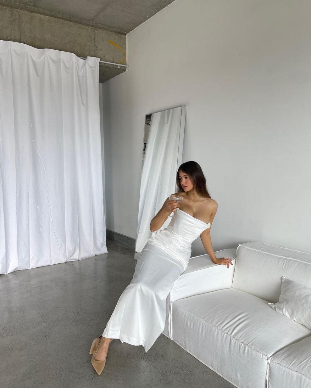 A woman in a strapless white floor-length dress and beige heels sits on a sofa arm, holding a glass in a concrete room.