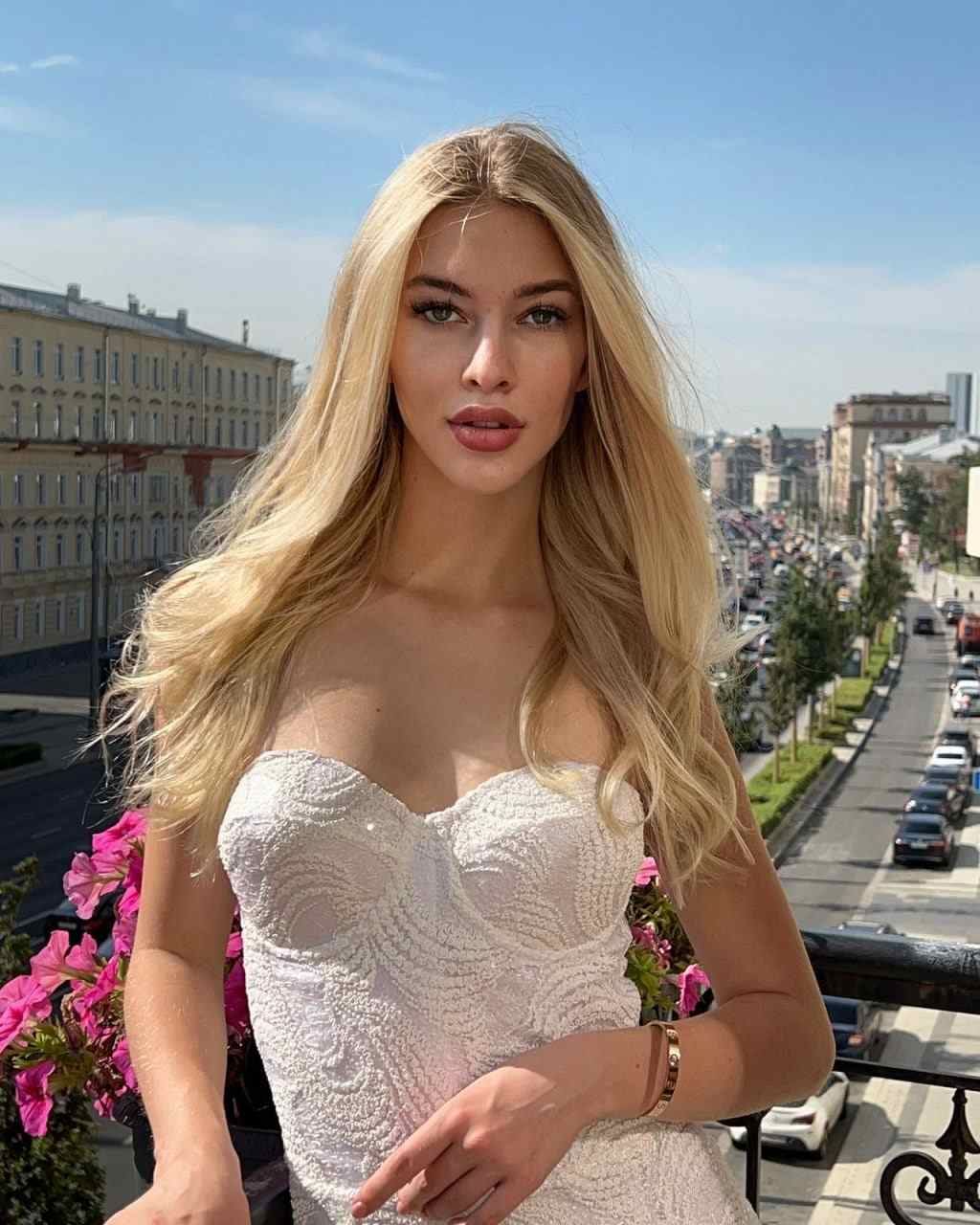Mari stands on a balcony in a white strapless lace dress, with city buildings and a blue sky in the background.