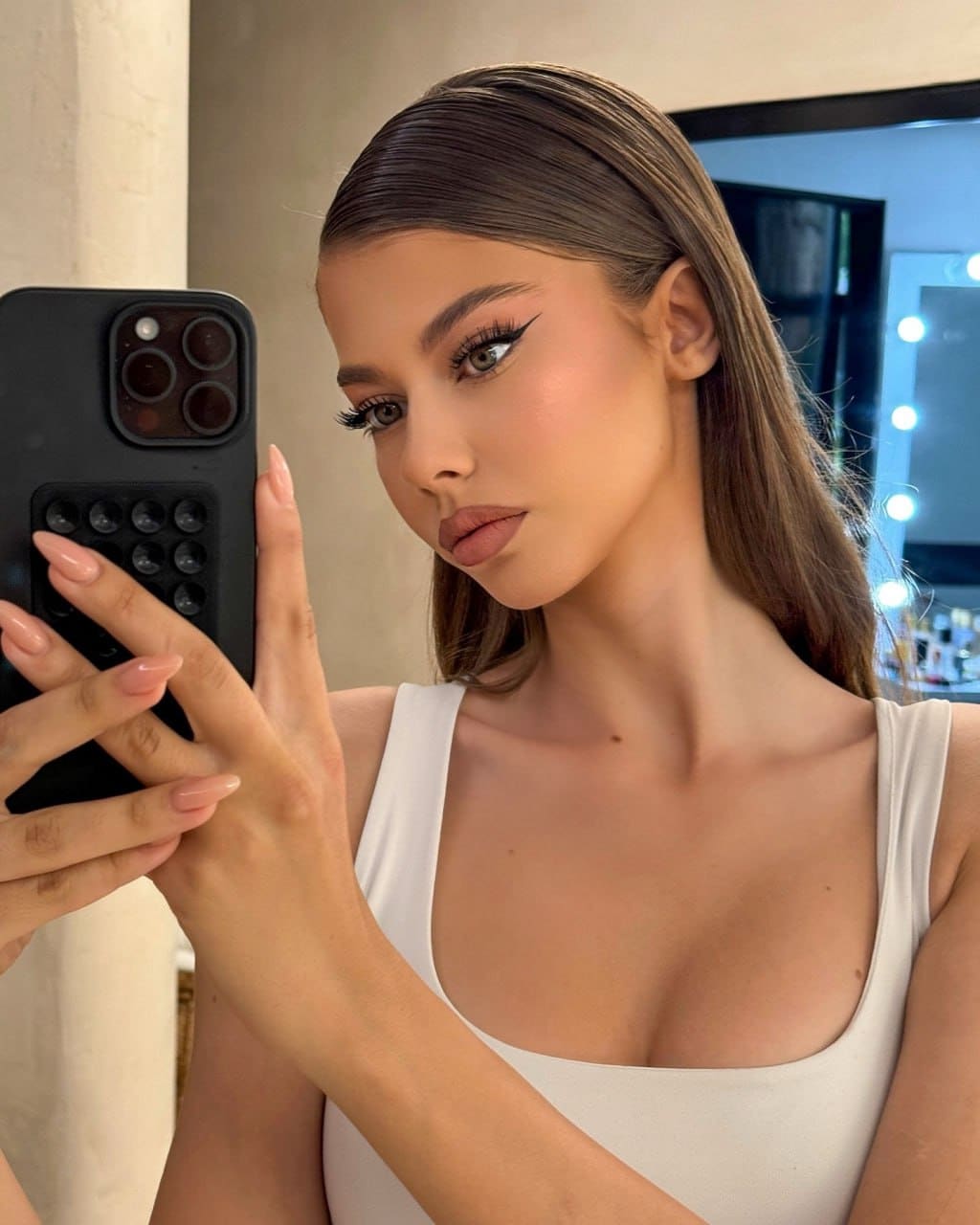 Clara takes a mirror selfie in a white sleeveless top with a scoop neck, holding a black smartphone in a room with a lit mirror.