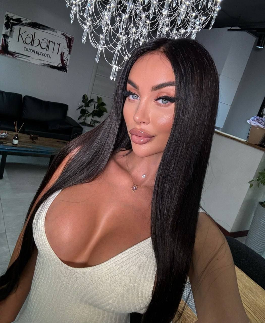 Poses indoors in a white ribbed top with a deep scoop neckline, beneath a crystal chandelier, with a black sofa and wall sign in the background.