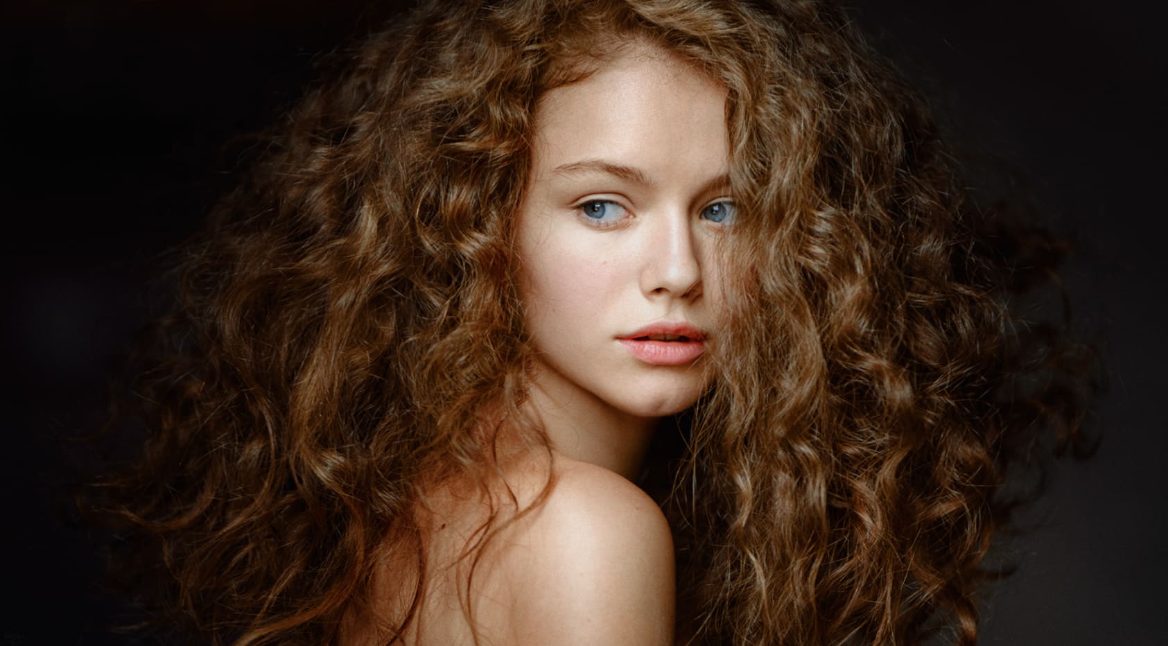 Close-up portrait of a person with voluminous curly brown hair and blue eyes, bare shoulders visible against a dark background.