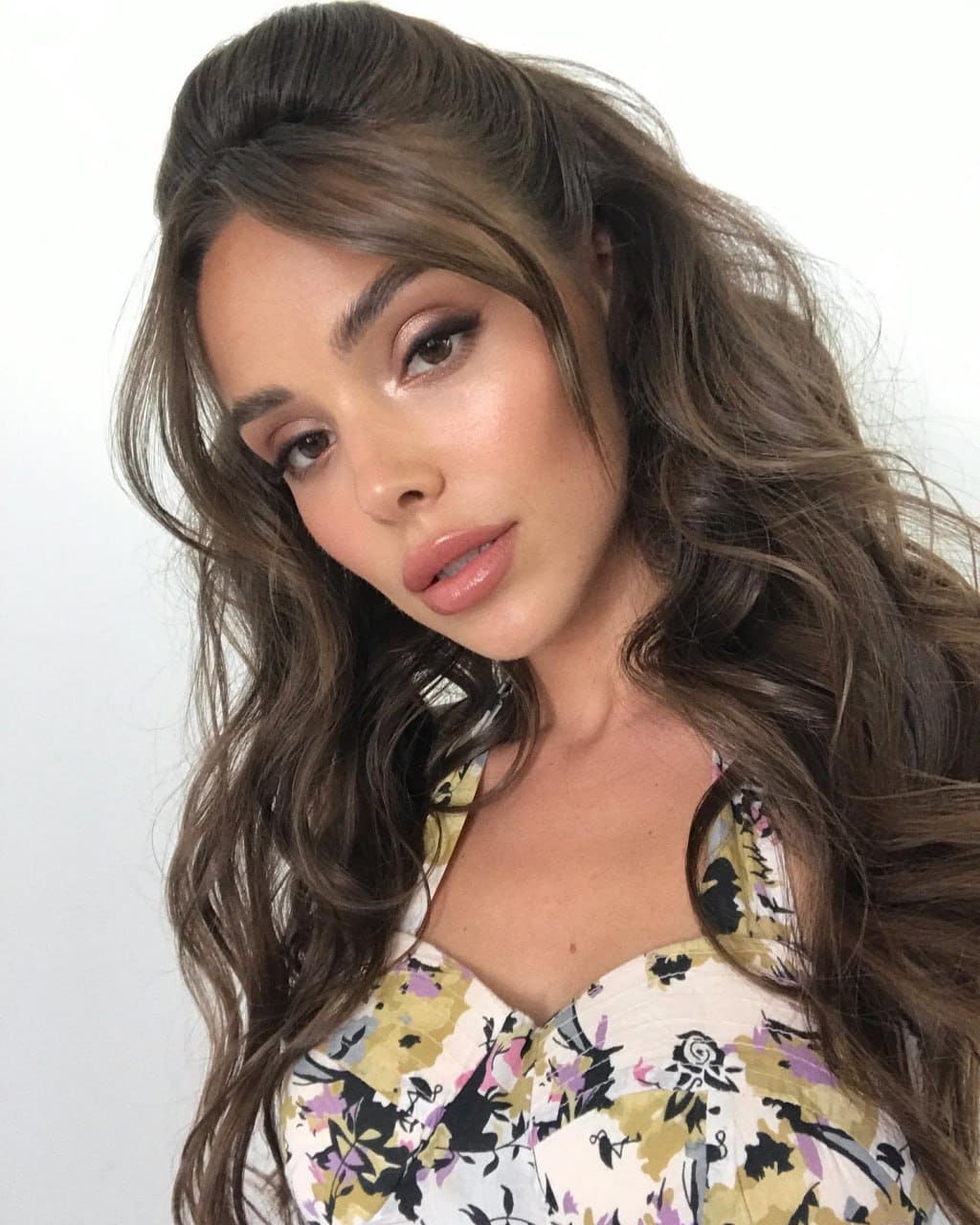 Anika with long wavy brown hair in a half-updo poses against a white background, wearing a floral top with a low neckline.