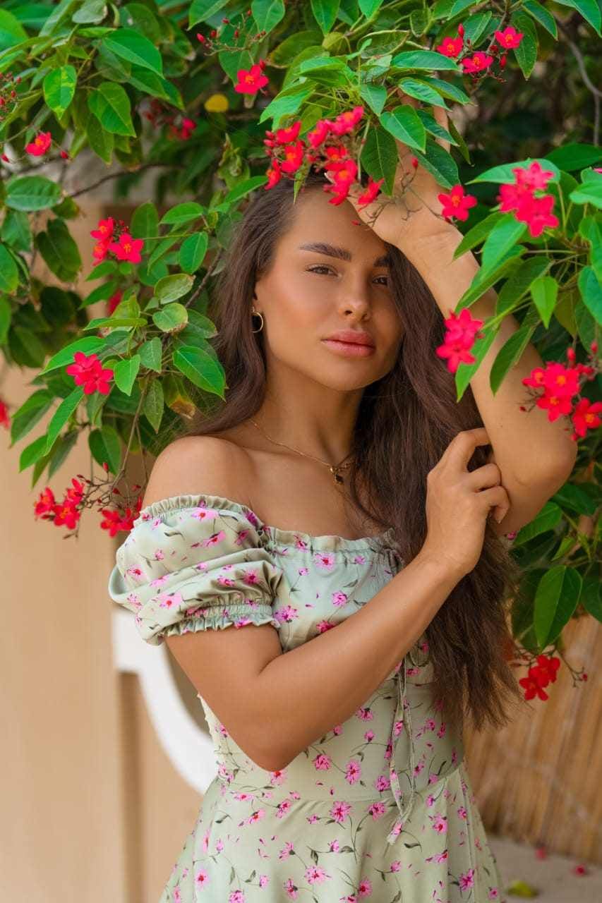 Lika stands among green leaves and pink flowers in a light green off-the-shoulder dress with a floral print, one hand raised to her hair.