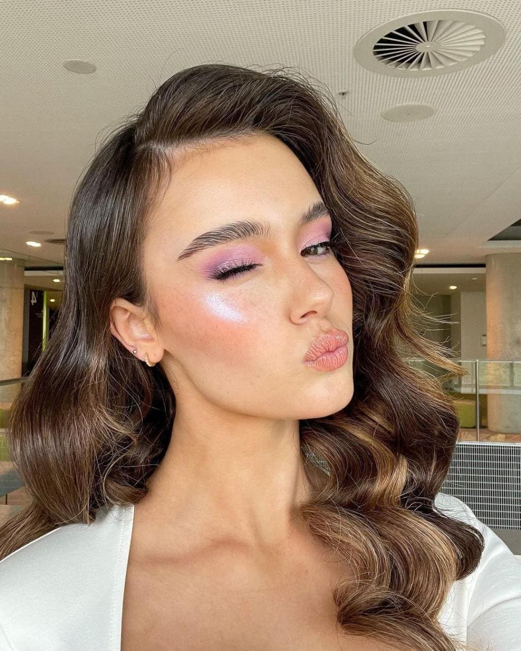 A woman with wavy brown hair and pink eyeshadow poses with pursed lips in a white top, indoors under a patterned ceiling.