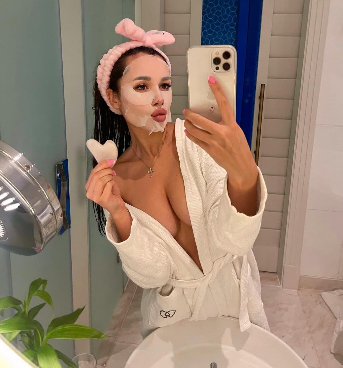 Woman in a bathroom wearing an open white bathrobe and facial mask, holding a phone and small object.