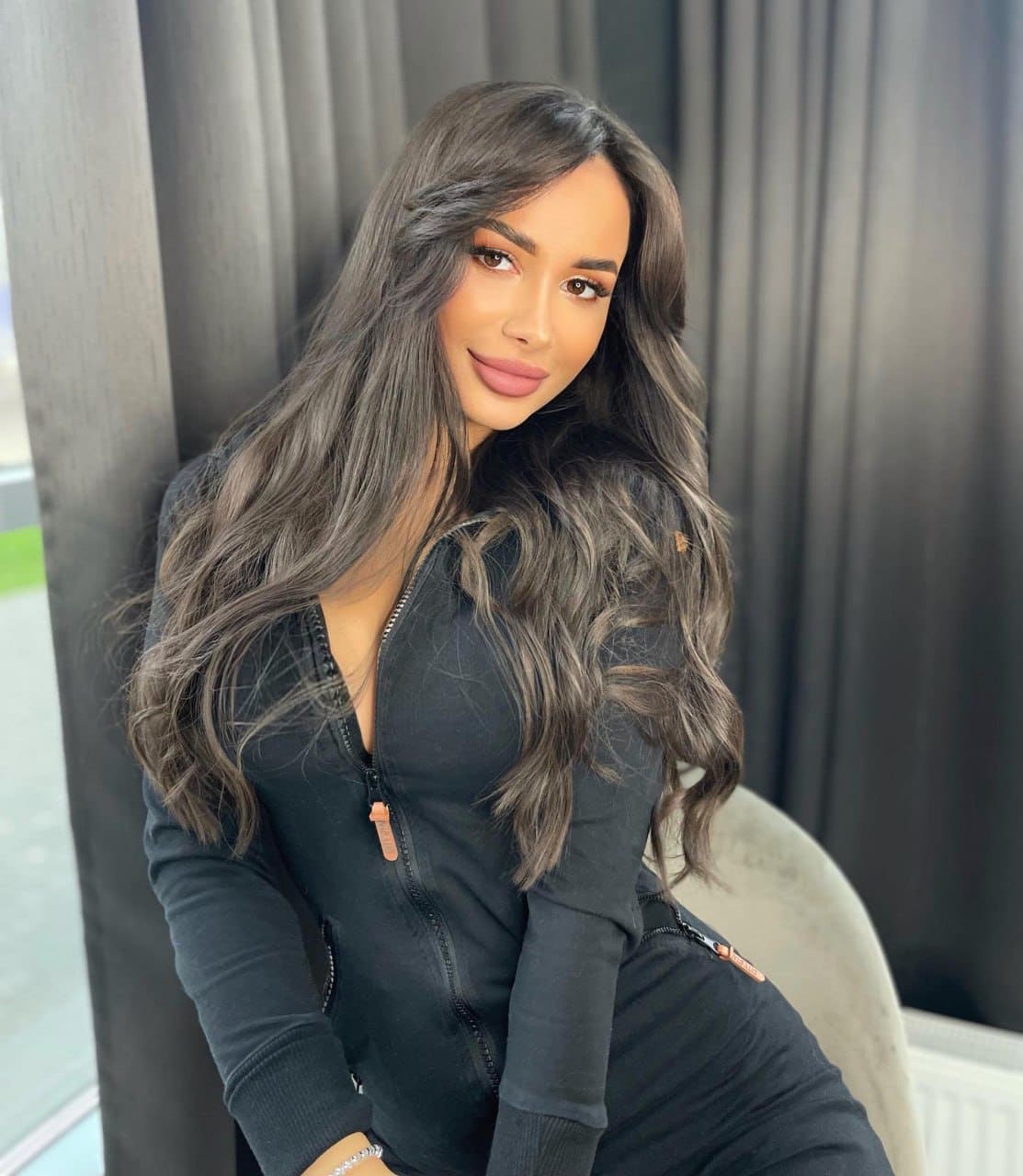 Polina sits in a light chair wearing a black zip-up top, against dark curtains and a window.
