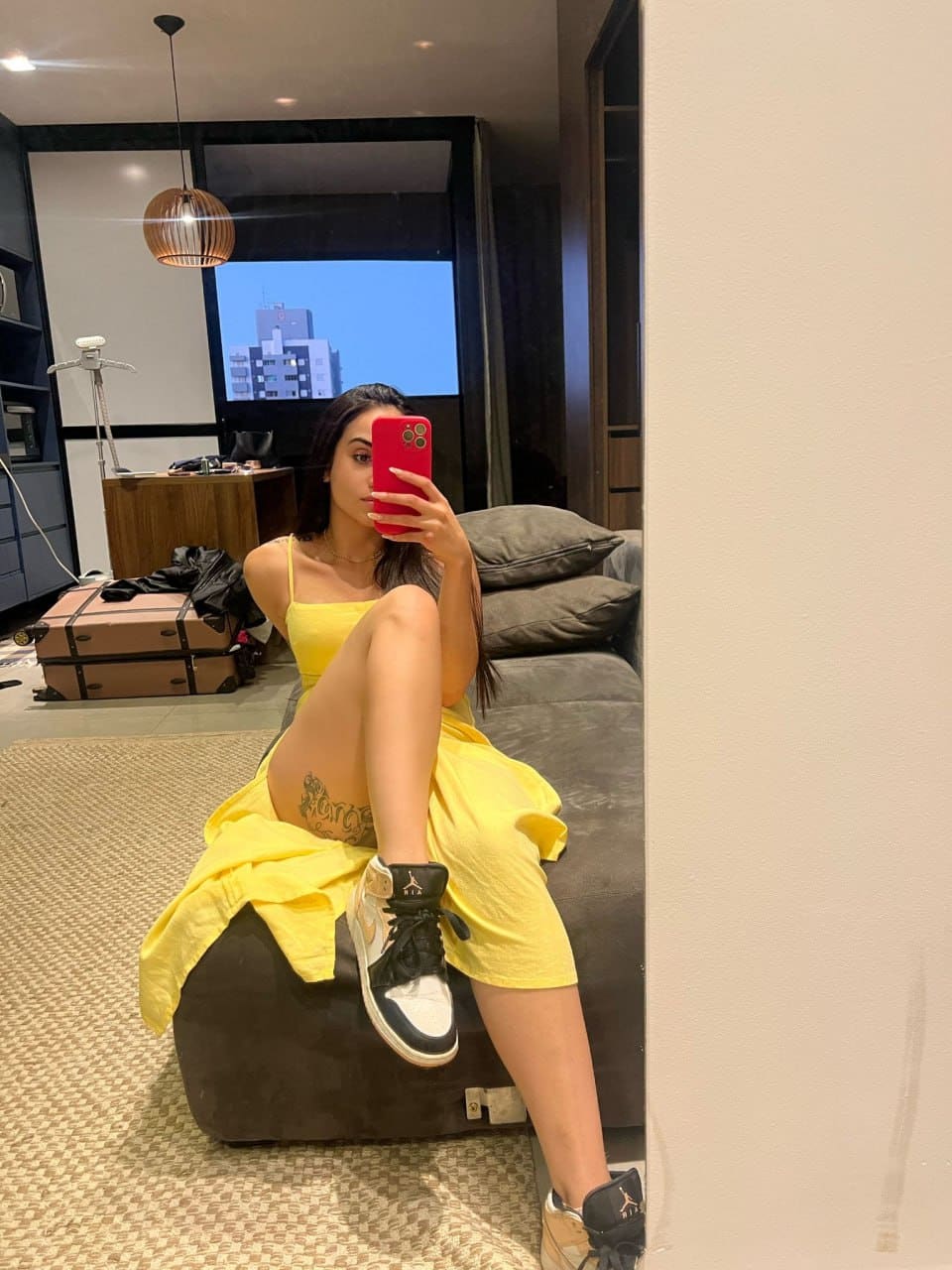 A woman sits on a dark ottoman in yellow clothing, holding a red phone to her face, one leg raised to show a thigh tattoo.