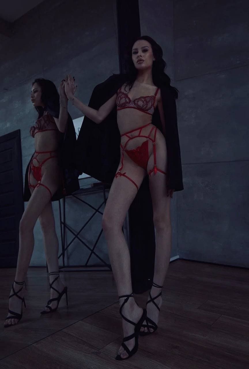 Standing in a dim concrete room, she wears red lingerie and a black coat, with a mirror reflecting her profile.