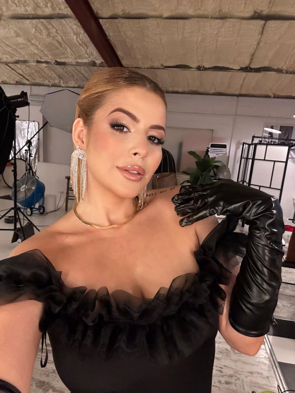 Woman in a black off-shoulder ruffled top, long black gloves, and earrings poses in a studio with lighting equipment visible.