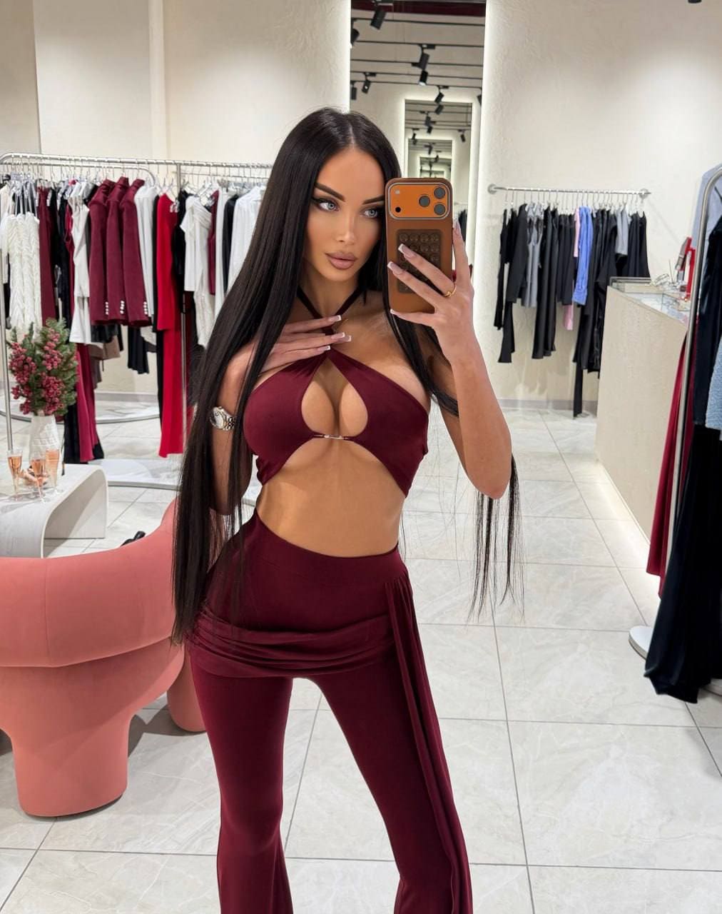 Mirror selfie in a clothing store wearing a maroon cut-out halter top and high-waisted pants, with clothes racks and a pink chair behind.