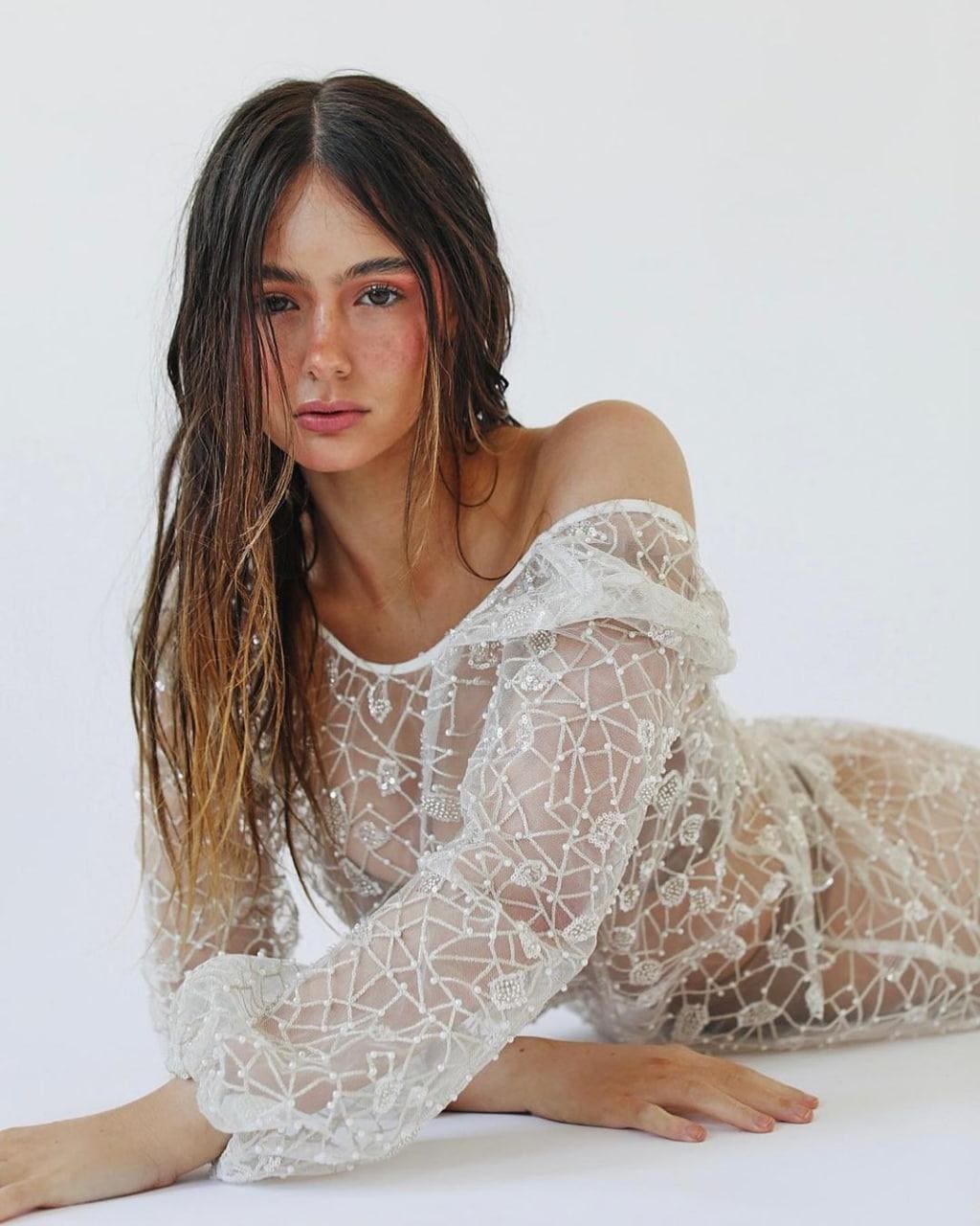 Holly lies on her side in a sheer white lace dress with long sleeves, wet dark hair against a plain white background.