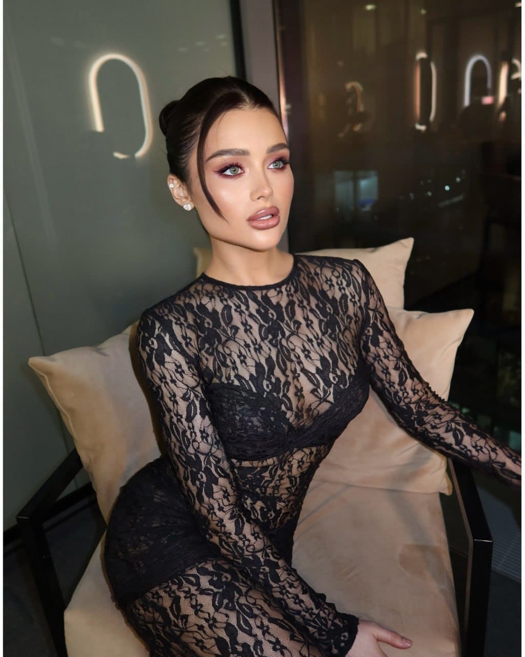 A woman sits on a beige chair wearing a black lace dress with a high neckline, in an indoor setting with soft lighting and a glass wall.