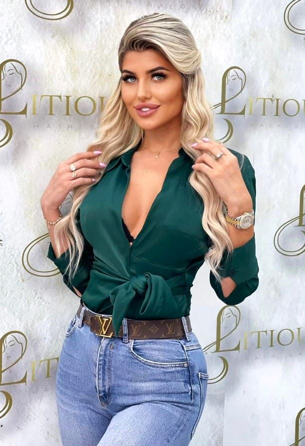 Natalia stands before a patterned backdrop in a dark green V-neck blouse and light blue jeans, holding her long wavy blonde hair with both hands.