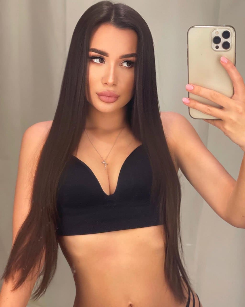 Mirror selfie of a woman in a black bralette and necklace, holding a phone against a plain light background.