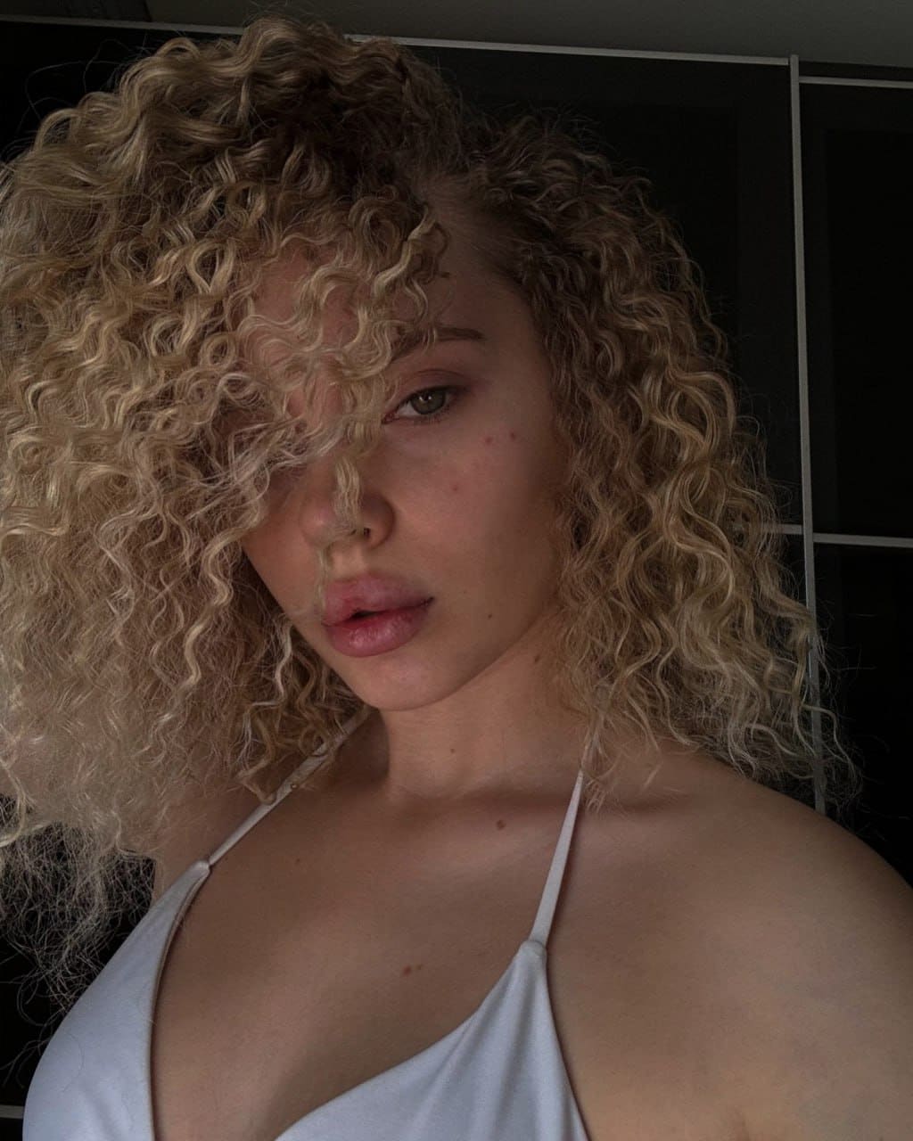 A close-up portrait shows voluminous curly blonde hair partially covering the face, wearing a white halter-neck top against a dark paneled background.