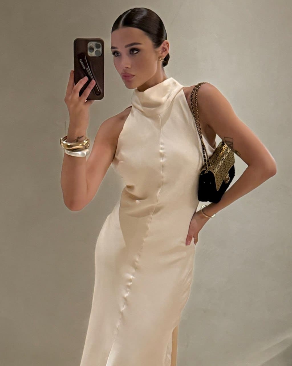 A woman takes a mirror selfie in a sleeveless cream satin dress with a high neckline, carrying a small chain-strap bag against a light wall.