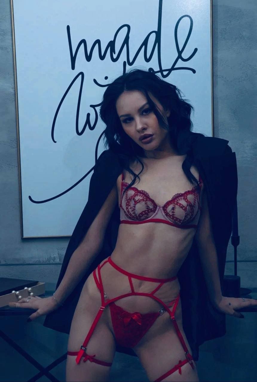 Carolina stands in red lace lingerie with a black jacket, posing before framed calligraphy artwork.