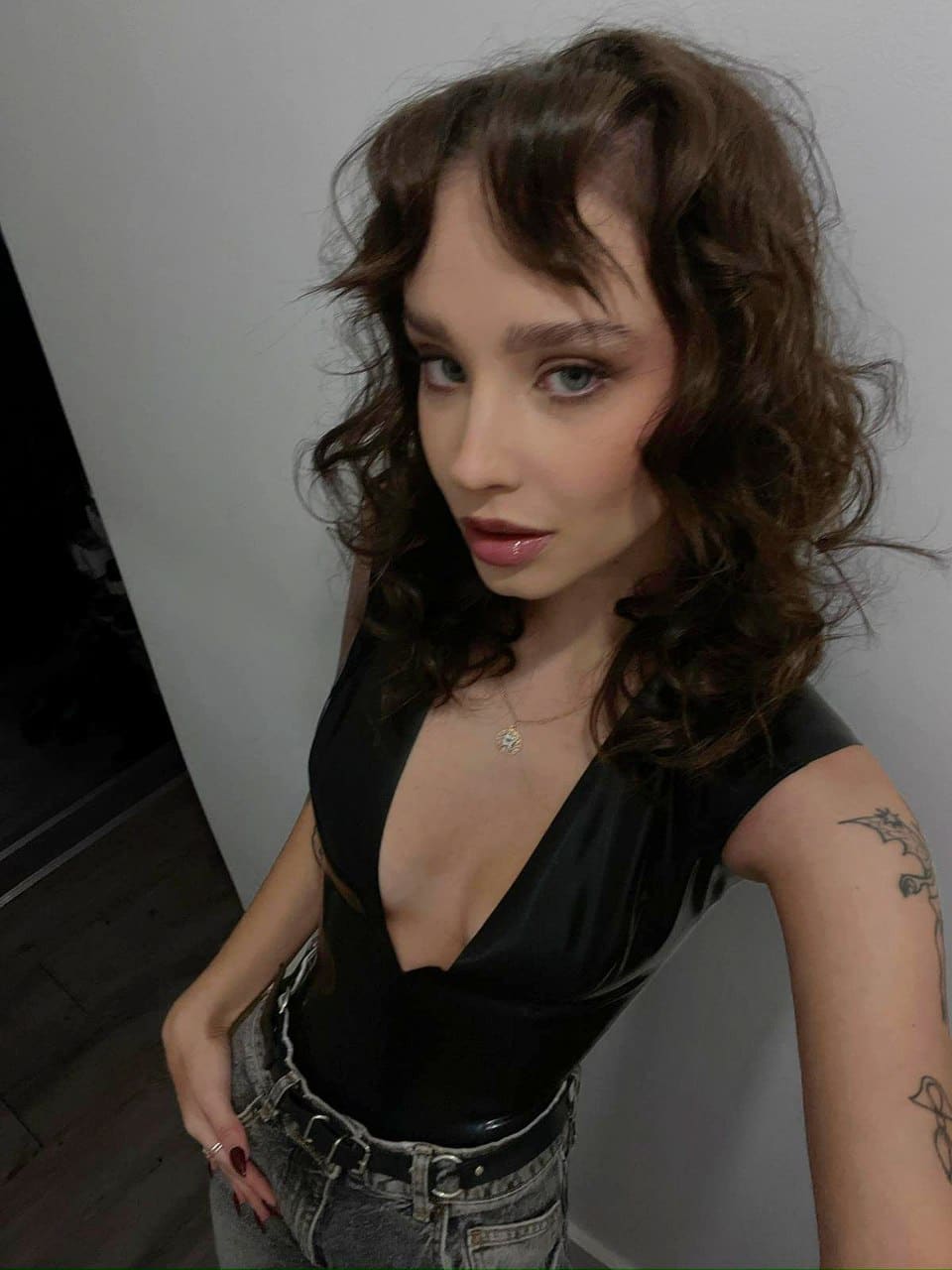 A woman with curly hair and bangs stands indoors in a black deep-V top and grey jeans, showing a tattoo on her left arm against a plain wall.