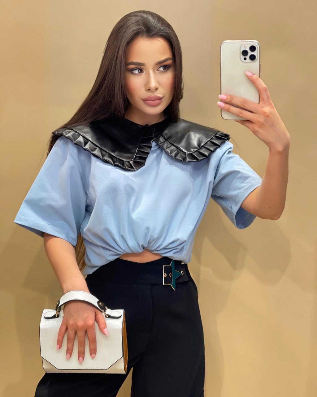Woman in a light blue blouse with black ruffled collar and star-buckle trousers, holding a white handbag and phone.