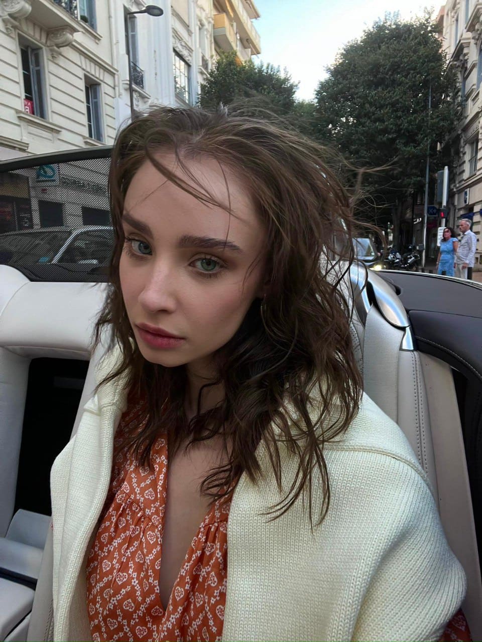 Daria sits in a car backseat wearing a white textured jacket and orange patterned blouse, with city buildings visible through the window behind her.